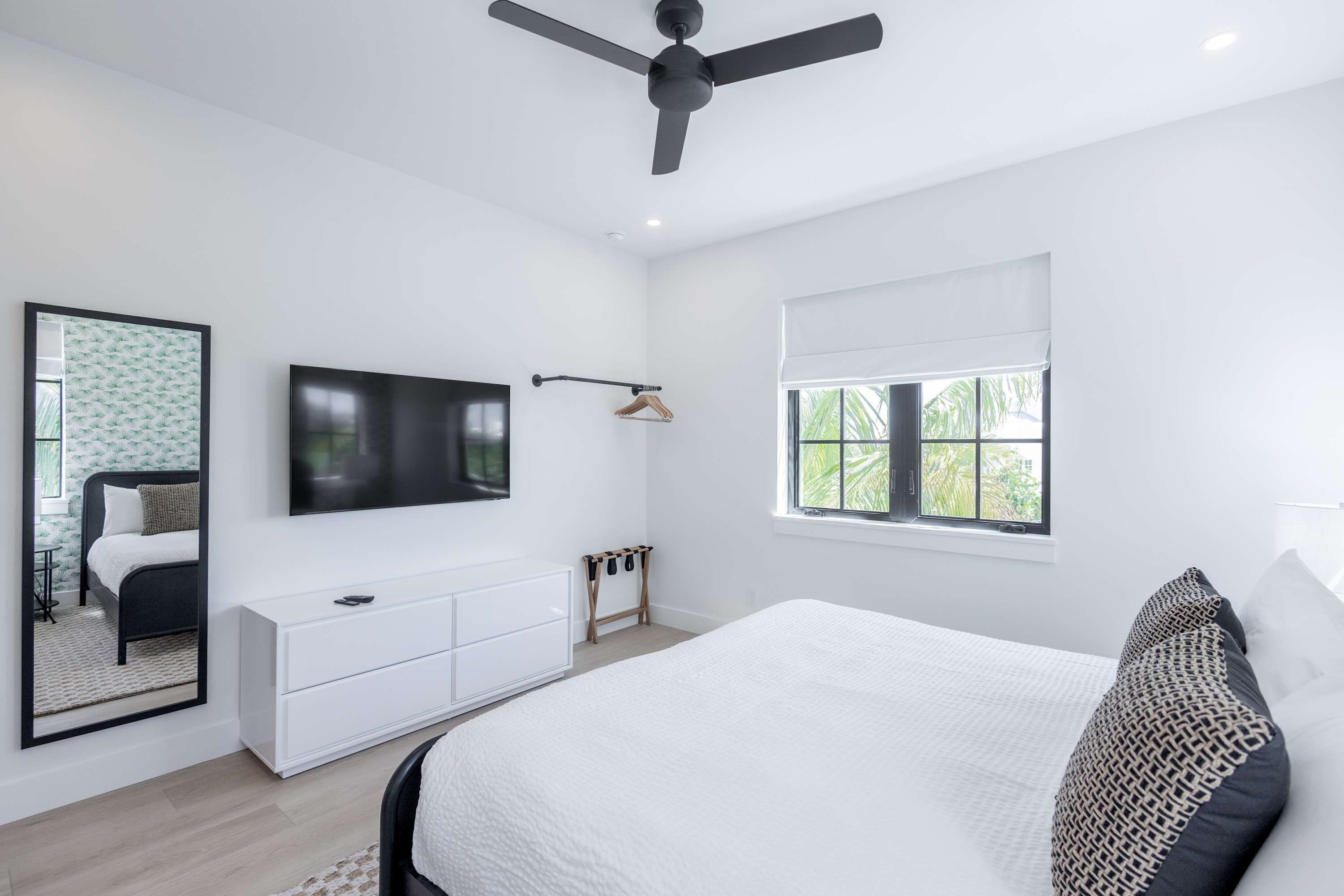 A modern bedroom with a large bed, a flat-screen TV mounted on the wall, a ceiling fan, a window with a view of greenery, and a bedside table with a lamp.