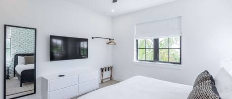 A modern bedroom with a large bed, a flat-screen TV mounted on the wall, a ceiling fan, a window with a view of greenery, and a bedside table with a lamp.