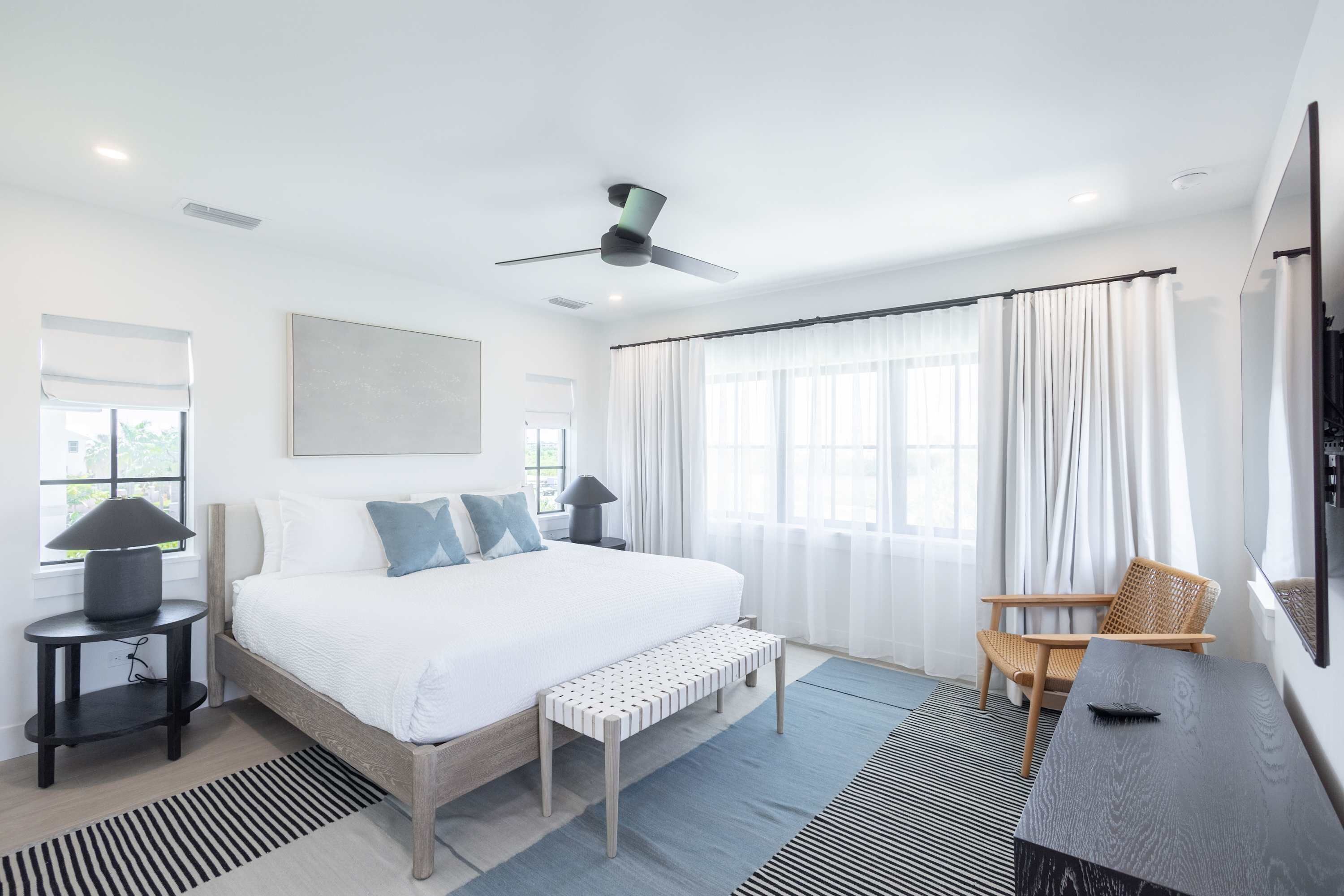 A modern bedroom with a large bed, a ceiling fan, a bedside table with a lamp, a bench, a chair, and a TV mounted on the wall.