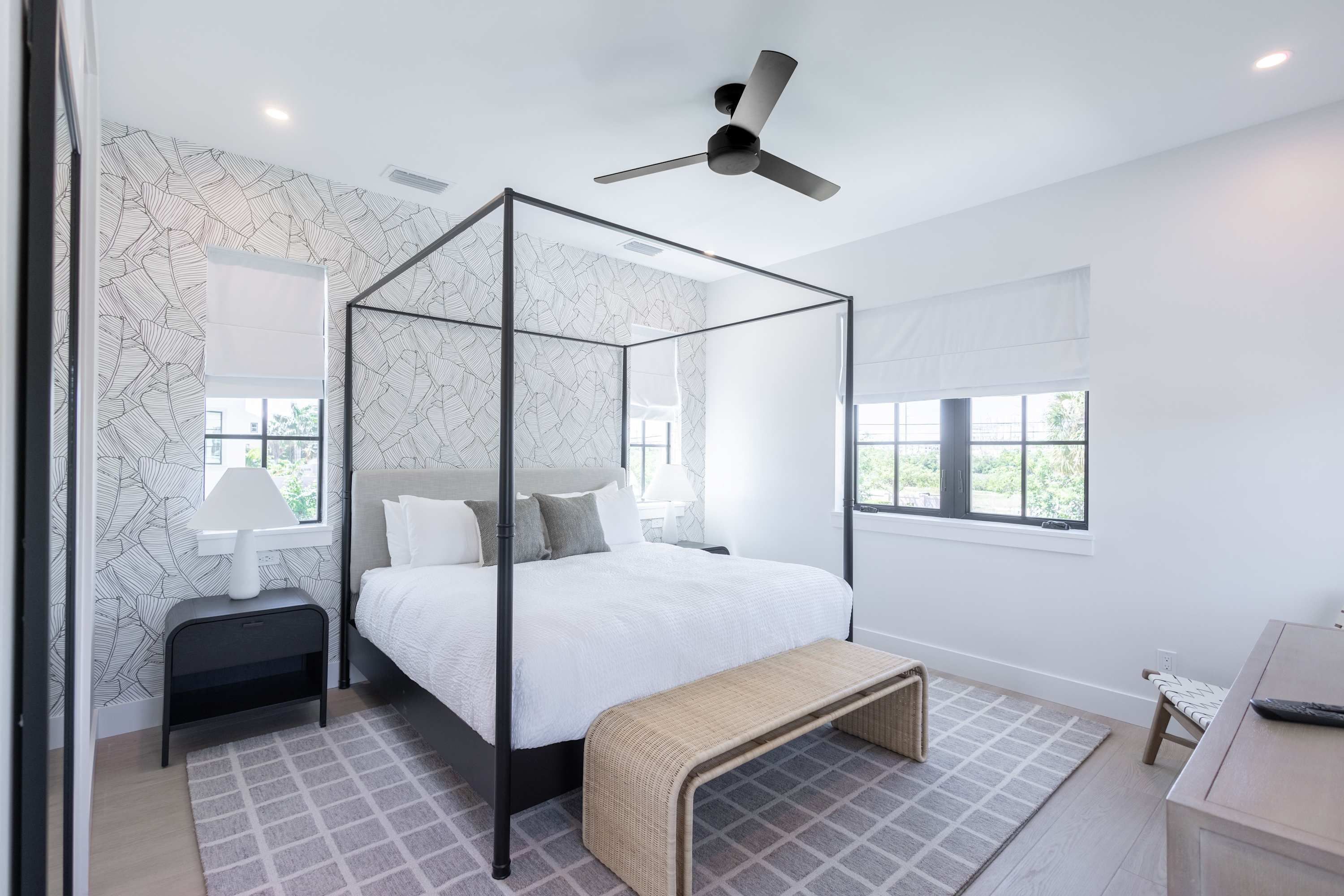 A modern bedroom with a four-poster bed, a ceiling fan, and a view of greenery through the window.
