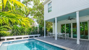 A swimming pool with a wooden deck, surrounded by lush greenery and a white building with a covered patio area. - NEW The Rigby House - Pvt Pool Amazing Location (Key West)