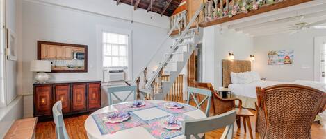 Una habitación acogedora y bien iluminada con techo de madera, un comedor con mesa y sillas redondas, una escalera con barandilla floral, y una cama con cabecera blanca.