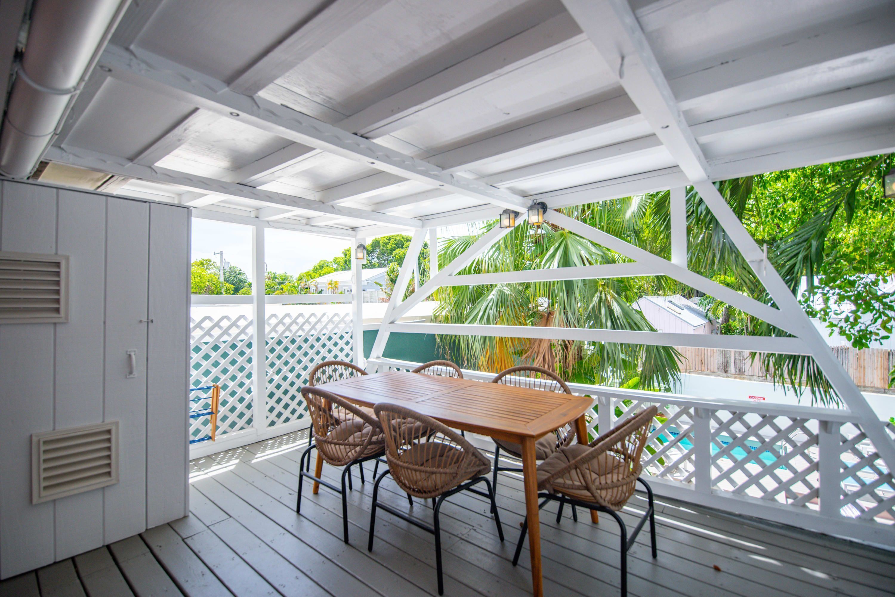 A covered outdoor area with a wooden table and wicker chairs, a white fence, and a view of a pool and greenery.