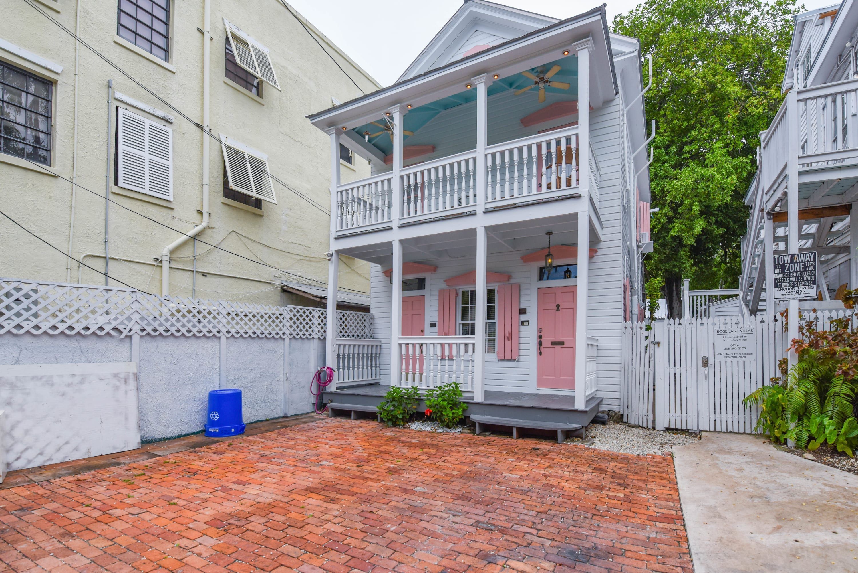 A two-story house with a balcony, a brick driveway, and a white picket fence.