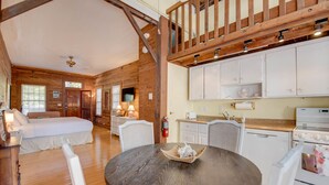 Una habitación acogedora con techo de madera, una mesa redonda con una cesta, muebles blancos y una zona de cocina con armarios y electrodomésticos blancos.
