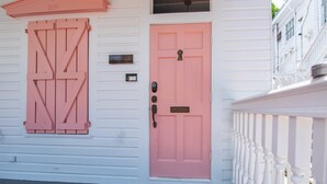Un edificio bianco con una porta e persiane rosa, una cassetta postale e una ringhiera bianca.
