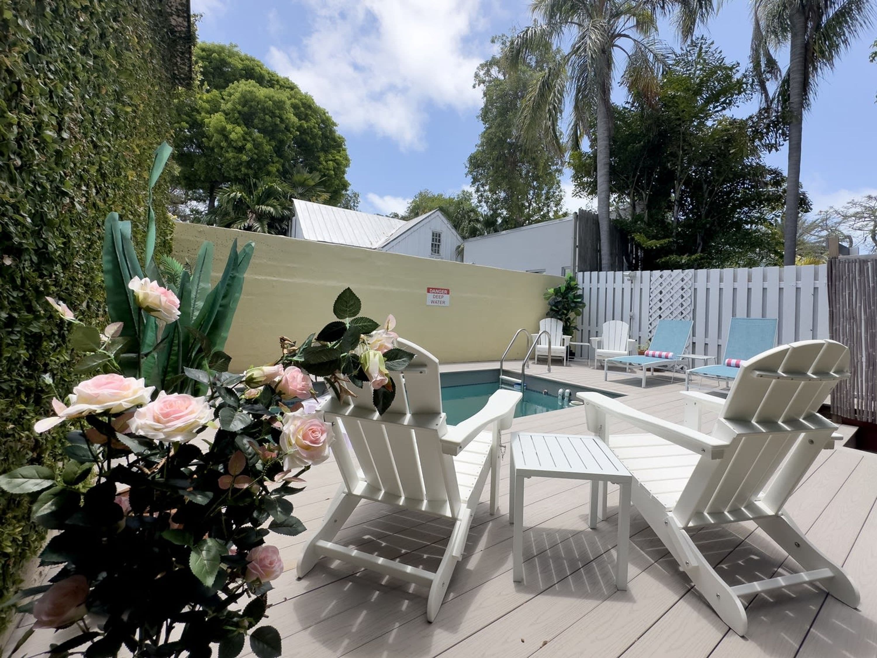 A poolside area with white lounge chairs and a small table, surrounded by a white fence and greenery.