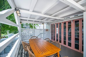 A covered outdoor seating area with a wooden table and wicker chairs, white lattice fence, and a pink door with glass panels. - New Rose Retreat - Pool Parking (Key West)