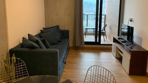 Een moderne woonkamer met een donkergrijze bank, een flatscreen televisie op een houten dressoir, en een balkon met uitzicht op de stad.