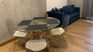 Een moderne woonkamer met een ronde glazen tafel, omringd door stoelen met een metalen frame, een donkerblauwe bank met kussens, en een betonmuur.