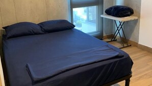 Een kamer met een enkel bed, een houten vloer, een raam met uitzicht op gebouwen en een klein tafeltje met een kussen erop.