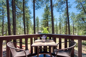 Una terraza de madera con una mesa y dos sillas de mimbre, con vistas a un bosque de altos pinos.
