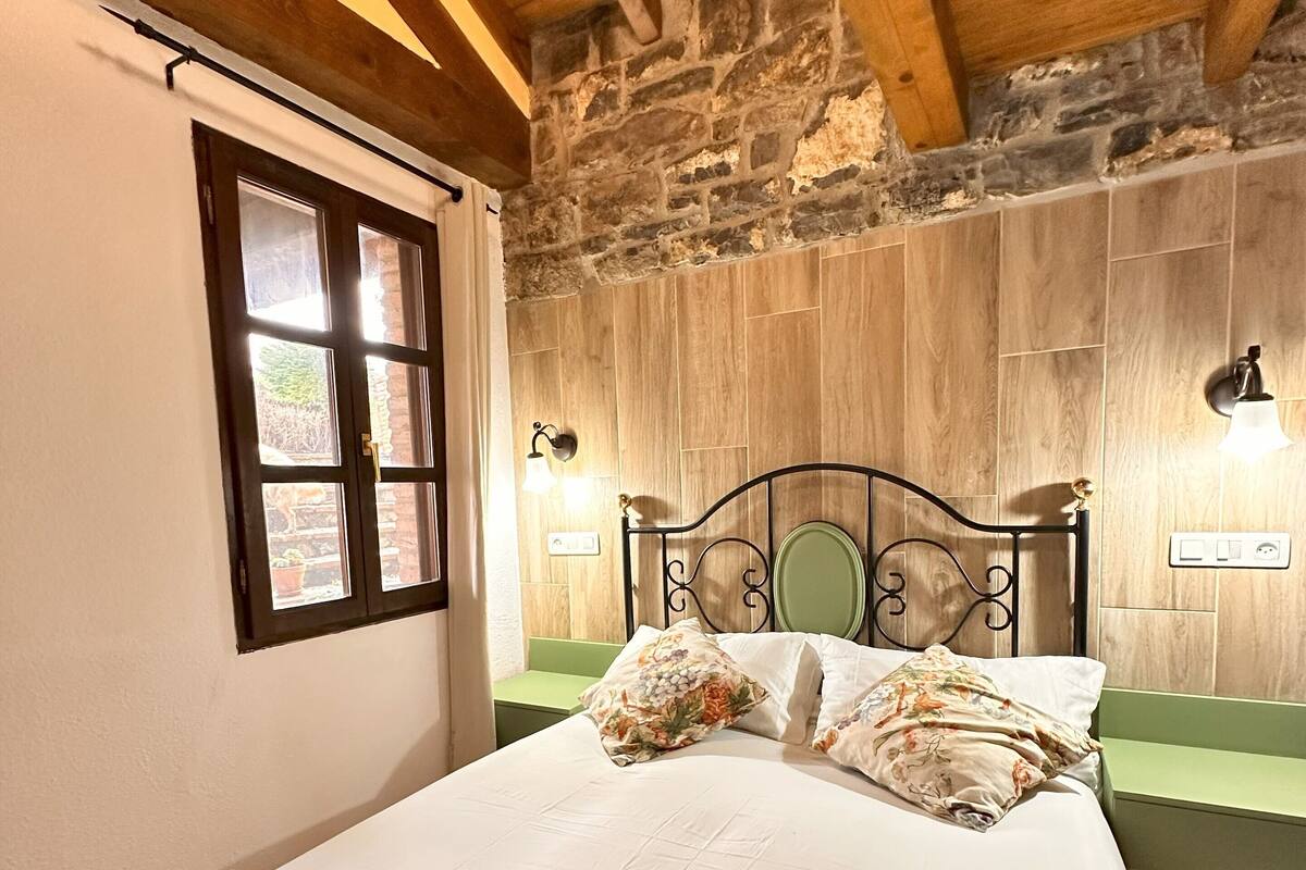 Un dormitorio con techo de madera, pared de piedra, cama con ropa de cama floral, un banco verde y una ventana con vistas.