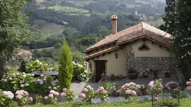 Una casa de piedra rústica con techo de tejas, rodeada de abundante vegetación y plantas en flor.