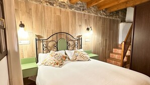 Un dormitorio con pared de piedra, vigas de madera en el techo, una cama con almohadones florales, una mesita de noche con lámpara y una escalera.
