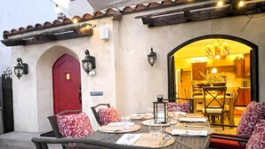 An outdoor dining area with a table set for two, surrounded by patterned chairs, a red door, and wall-mounted lights. - Steps from the sand, well-stocked, clean, family beach home. AC & easy pkg. (Long Beach)
