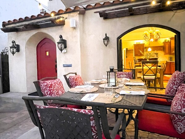 An outdoor dining area with a table set for two, surrounded by patterned chairs, a red door, and wall-mounted lights.