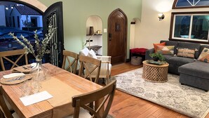 A dining area with a wooden table, chairs, and a vase with flowers. There is a sofa and a rug in the living area. - Steps from the sand, well-stocked, clean, family beach home. AC & easy pkg. (Long Beach)