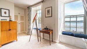 Een kamer met een houten kast, een telescoop, een klein tafeltje met decoratieve objecten, een raam met uitzicht op zee en een ingelijst portret aan de muur.