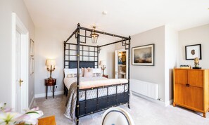 Una cama con dosel de estructura oscura y cabecera con estampado geométrico. Hay una mesita de noche con lámpara y un espejo en la pared. Un armario de madera se encuentra a la derecha de la cama.