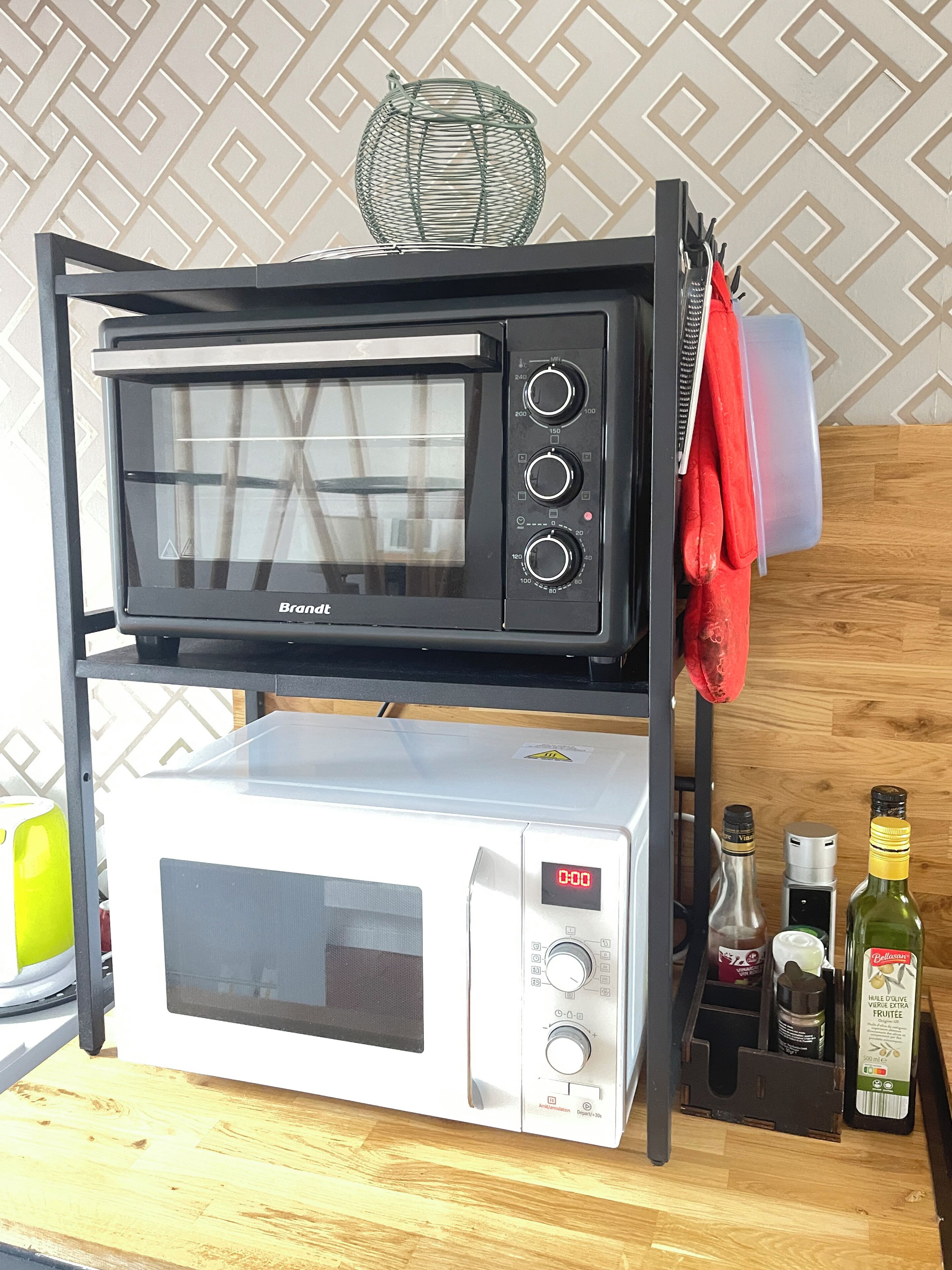 Uma cozinha com micro-ondas e forno elétrico empilhados em uma prateleira, uma toalha vermelha pendurada na prateleira, e uma garrafa de azeite no balcão.