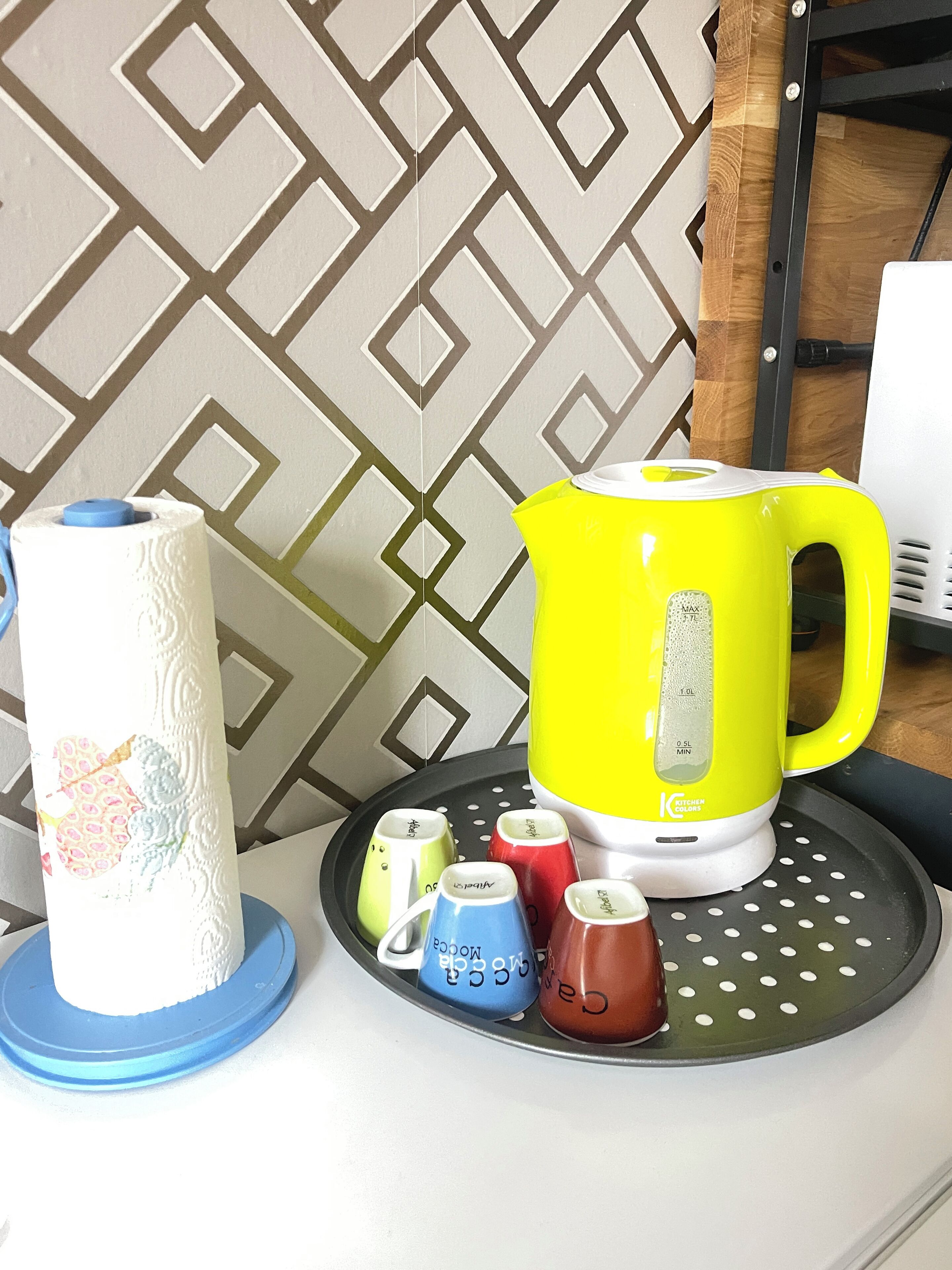 A kitchen area with a yellow kettle, a roll of paper towels, and colorful mugs on a tray.