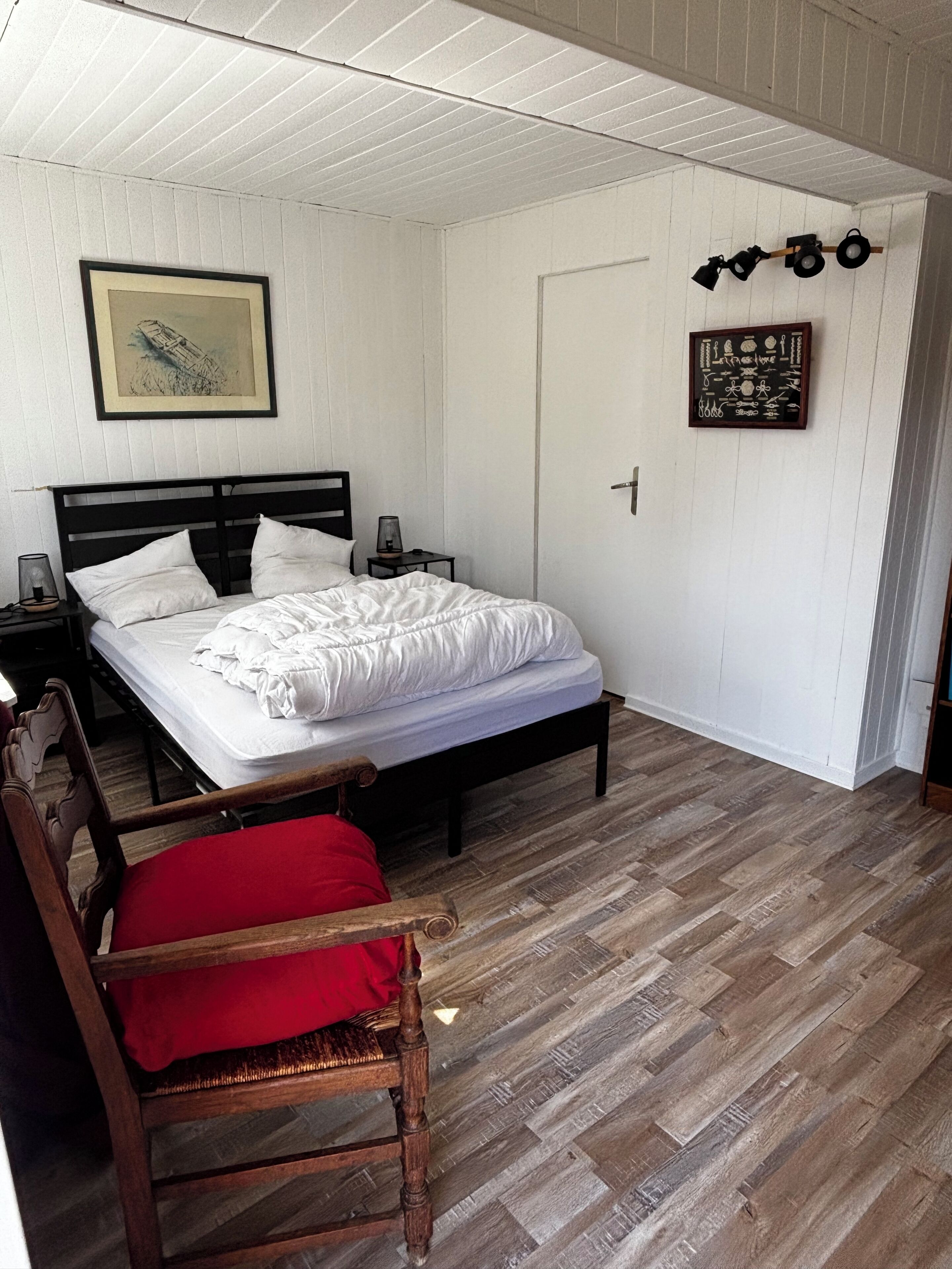 Een slaapkamer met een bed, een stoel, een nachtkastje, een aan de muur bevestigd schilderij en een deur.