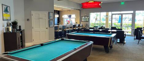 A pool hall with multiple pool tables, a bar area, and a view of the outdoors through large windows.