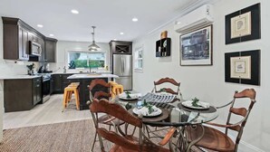 A modern kitchen with dark cabinetry, a central island, and a dining area with a glass table and metal chairs. - Battery Place (Savannah)