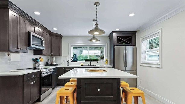A modern kitchen with dark cabinets, a central island, yellow stools, stainless steel appliances, and a large window. - Battery Place (Savannah)