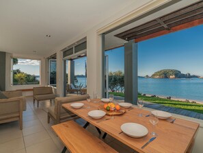 A dining area with a view of the sea, furnished with wooden tables and chairs, and a bench. - Hahei Oceanfront - Hahei Holiday Home (Hahei)