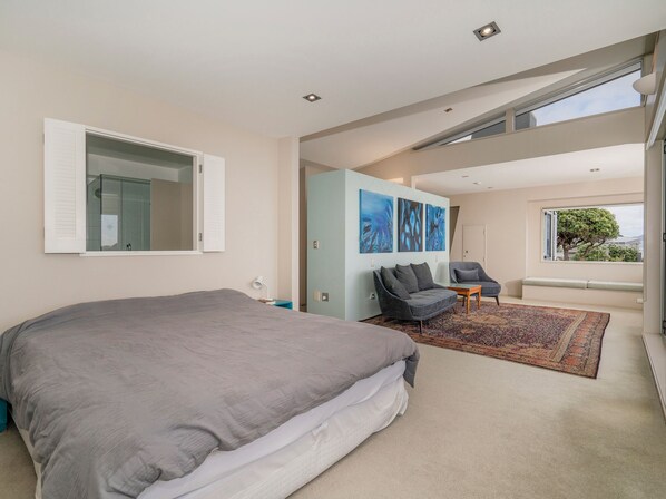 A modern bedroom with a large bed, a seating area with a sofa and chairs, and a view of the outdoors through a large window. - Hahei Oceanfront - Hahei Holiday Home (Hahei)