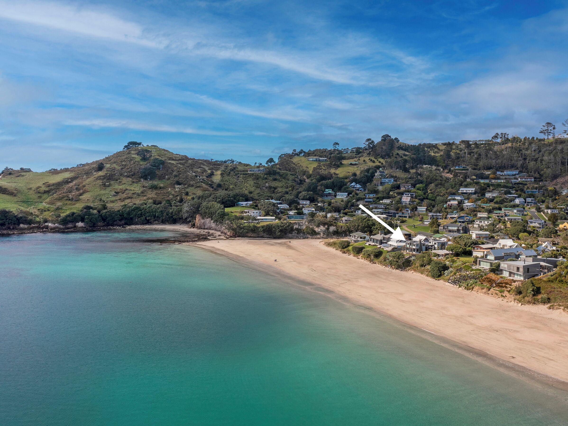A coastal area with a sandy beach, clear turquoise waters, and a hillside with residential buildings.