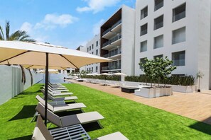 Et moderne udendørs opholdsområde med liggestole og parasoller, en pool samt en bygning med altaner.