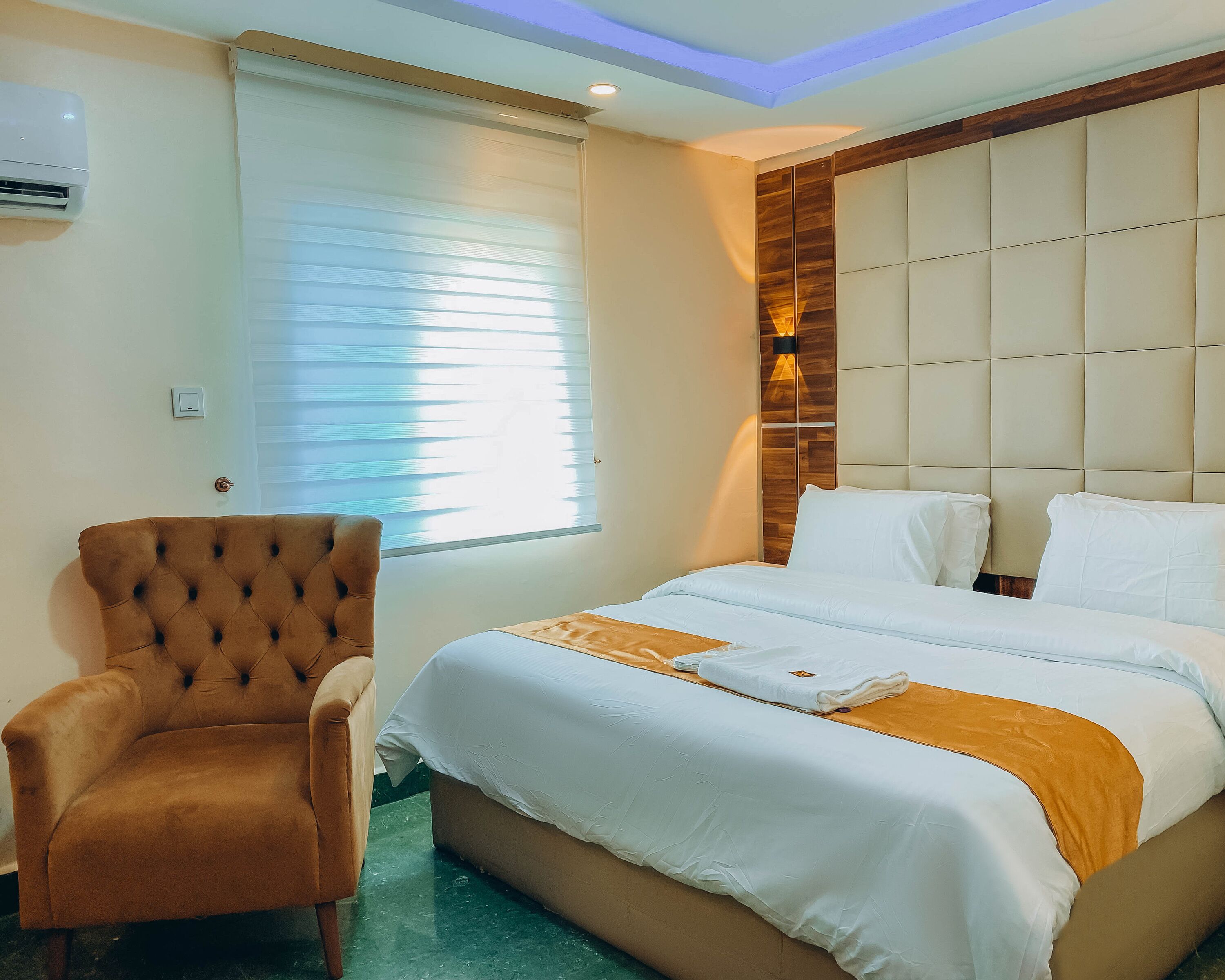 A hotel room with a bed, a tufted armchair, a window with blinds, and a wall-mounted air conditioner.