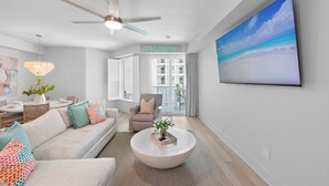 Een moderne woonkamer met een witte bank, een ronde salontafel, een plafondventilator en een groot televisiescherm dat een strandscène toont.
