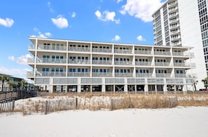 Um hotel de vários andares com varandas, uma praia arenosa em frente e um céu azul límpido.
