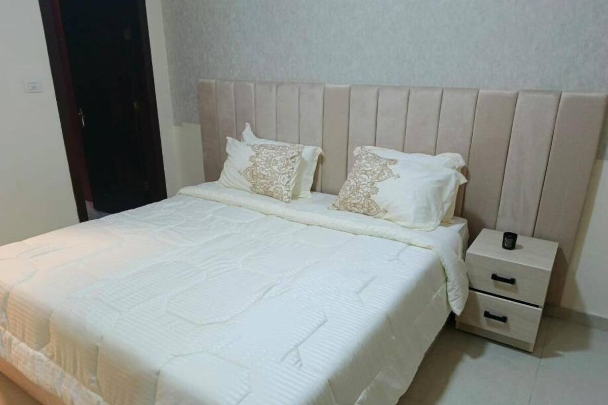 A neatly made bed with a quilted cover and two decorative pillows, a bedside table with a drawer, and a plain wall in a hotel room.