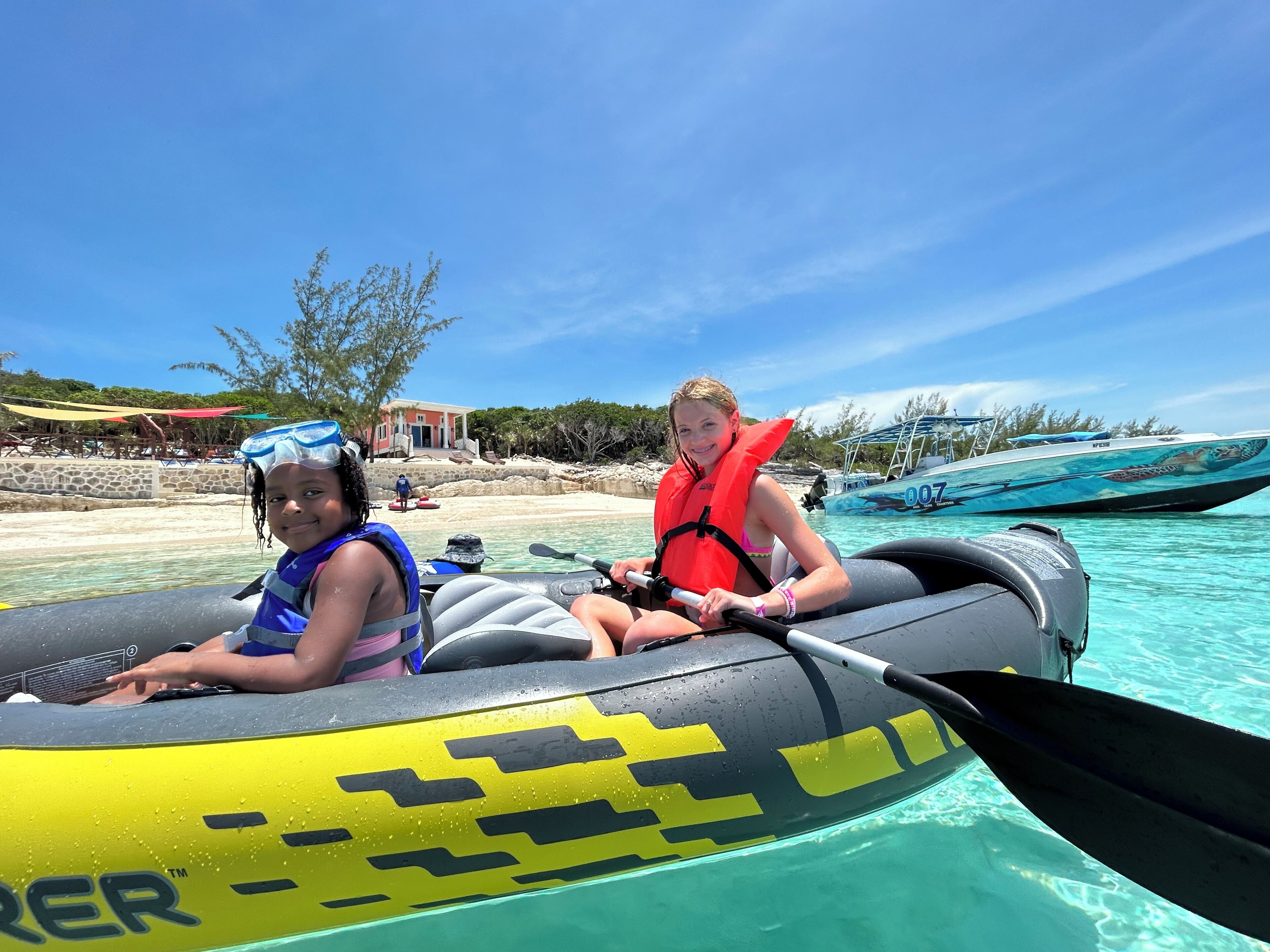 Deux personnes sur un radeau gonflable, avec une vue dégagée sur une plage et des bateaux en arrière-plan.