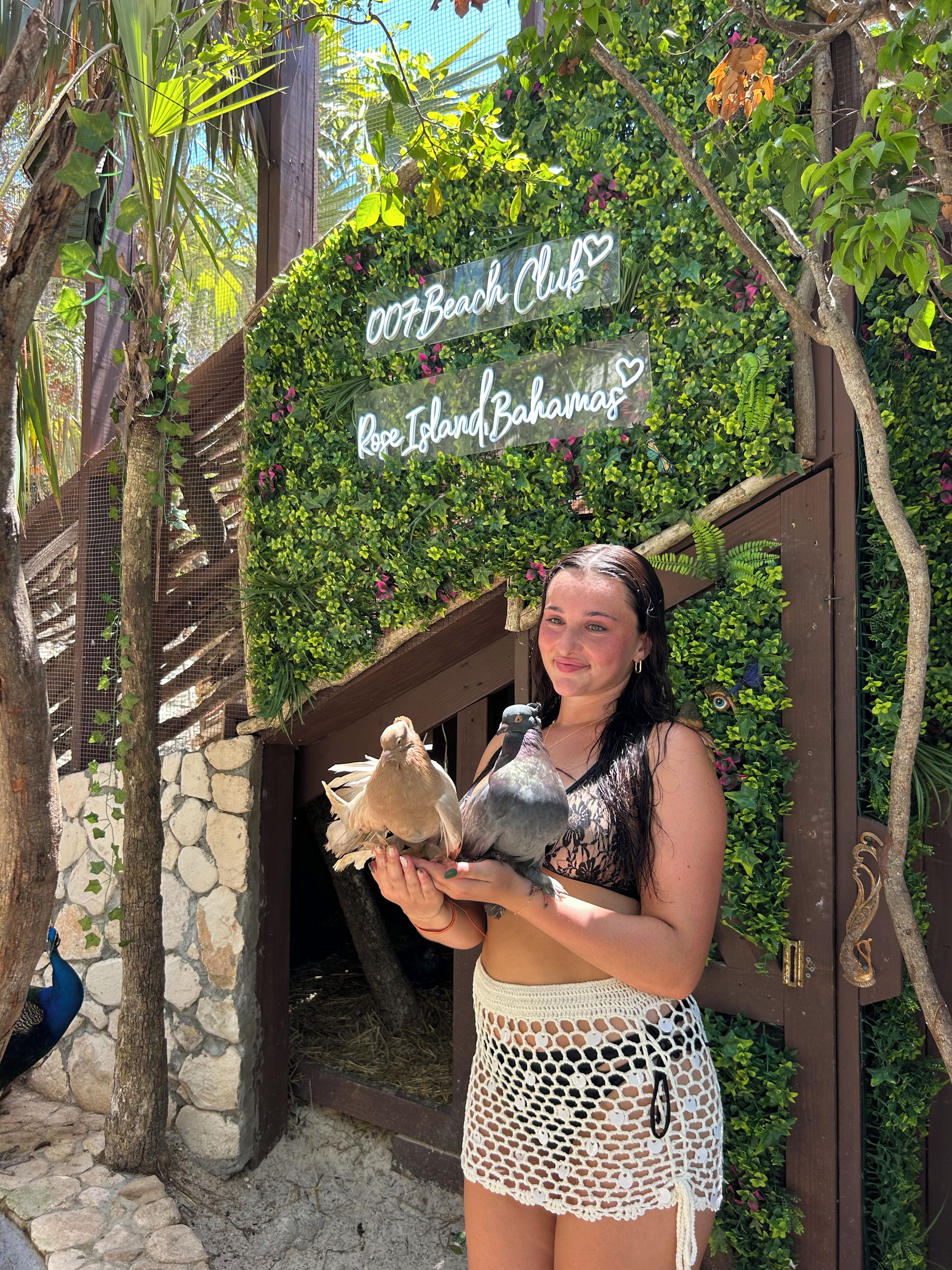 A person holding two pigeons in front of a sign that reads 'Beach Club Rose Island Bahamas.'