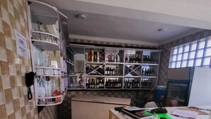 A bar area with shelves of bottles and glasses, a counter with a phone and papers, and a patterned floor.
