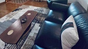A living room with a black leather sofa, a wooden coffee table, and a patterned rug. There is a window with curtains, a door, and a wall clock. - Green Acres (Minford)
