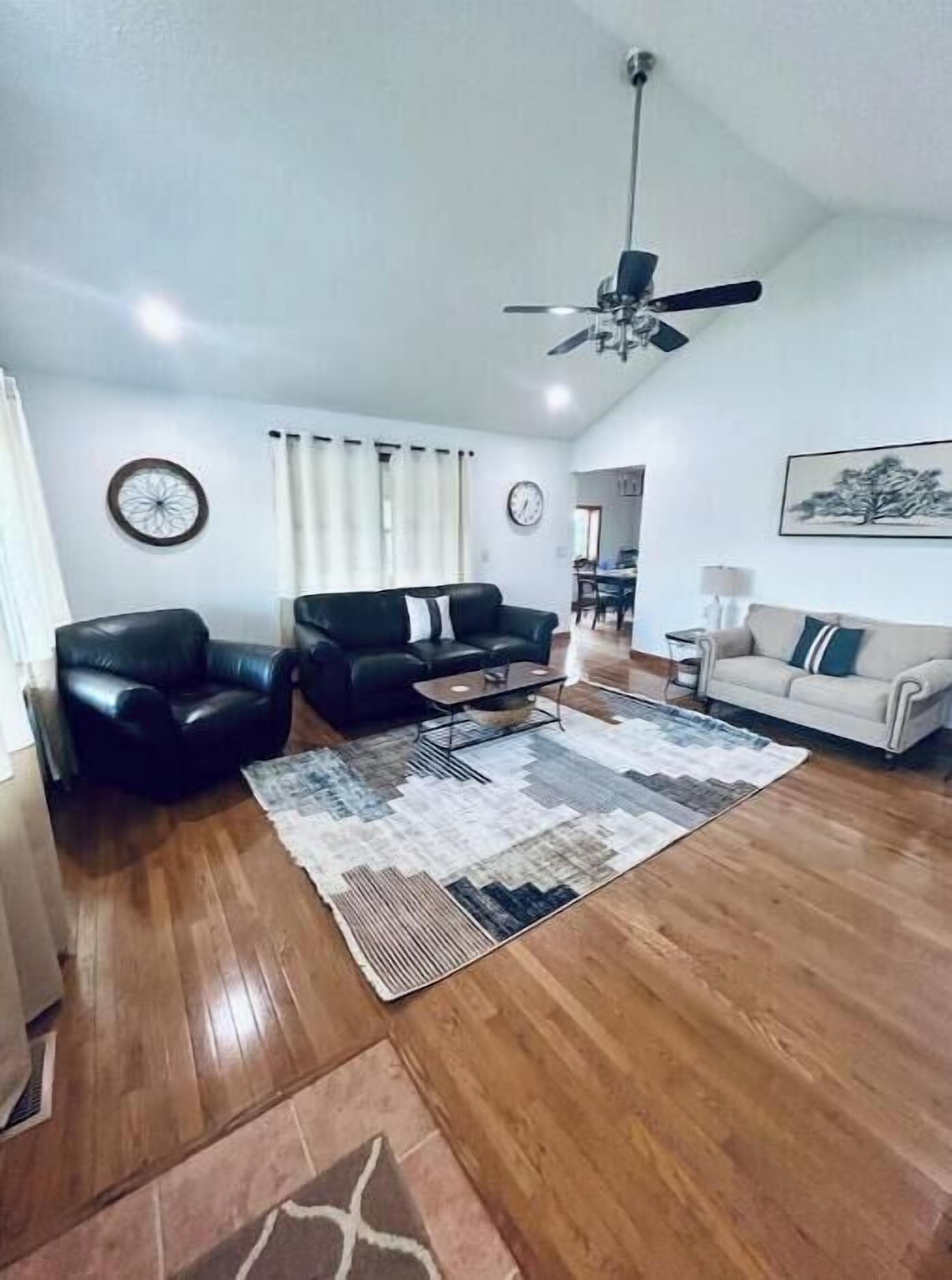 A spacious living room with a black leather sofa, a beige sofa, and a coffee table. There is a ceiling fan, a clock, and a painting on the wall.