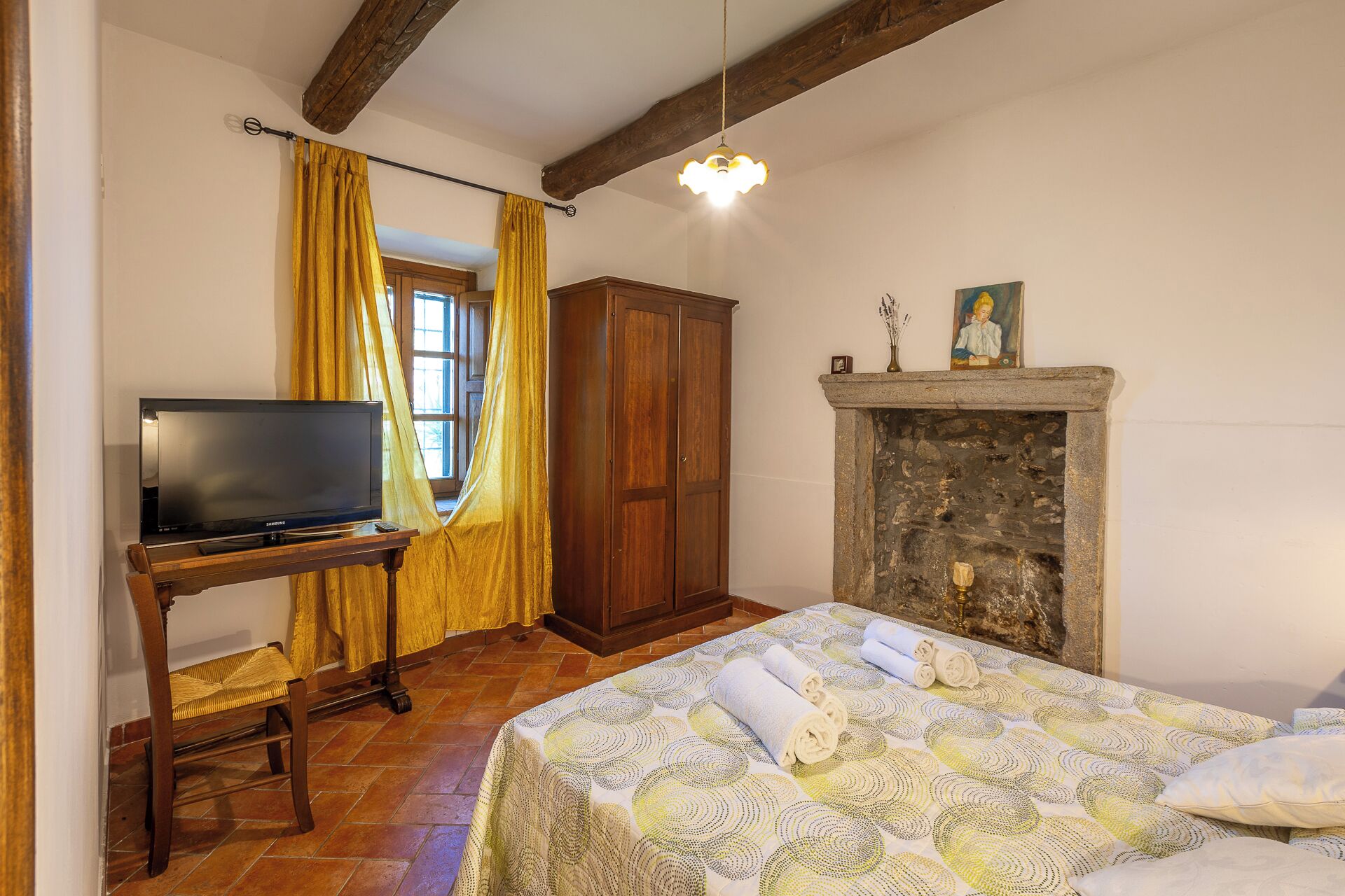 Quarto com cama, televisão sobre suporte de madeira, guarda-roupa, lareira e janela com cortinas.