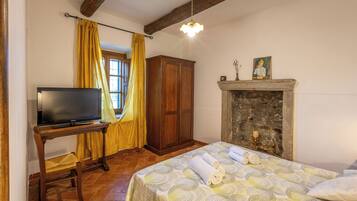 Quarto com cama, televisão sobre suporte de madeira, guarda-roupa, lareira e janela com cortinas.