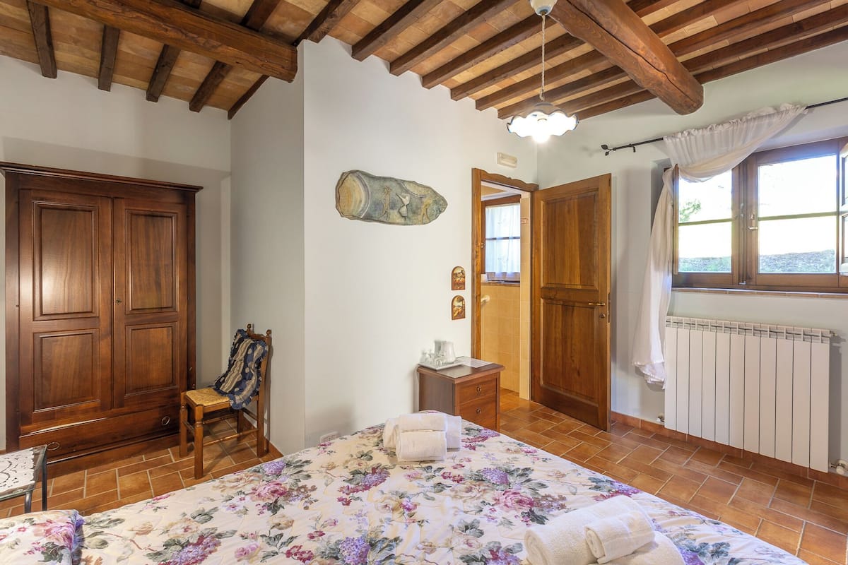 A bedroom with a wooden ceiling, a bed with floral bedding, a wooden wardrobe, a chair with a blanket, a small table with a lamp, and a window with curtains.