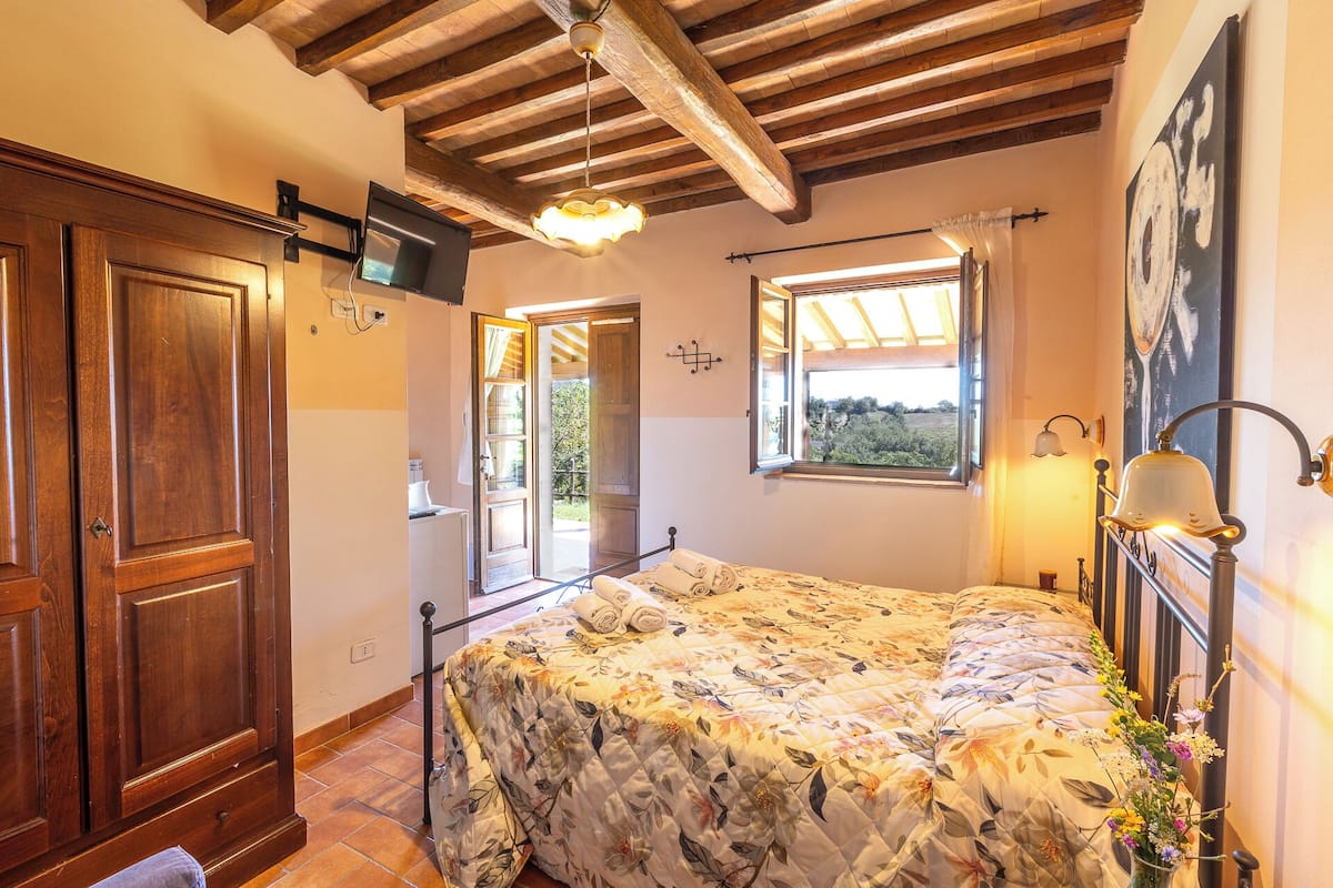 A bedroom with a bed, wooden ceiling beams, a window with curtains, and a wall-mounted TV.