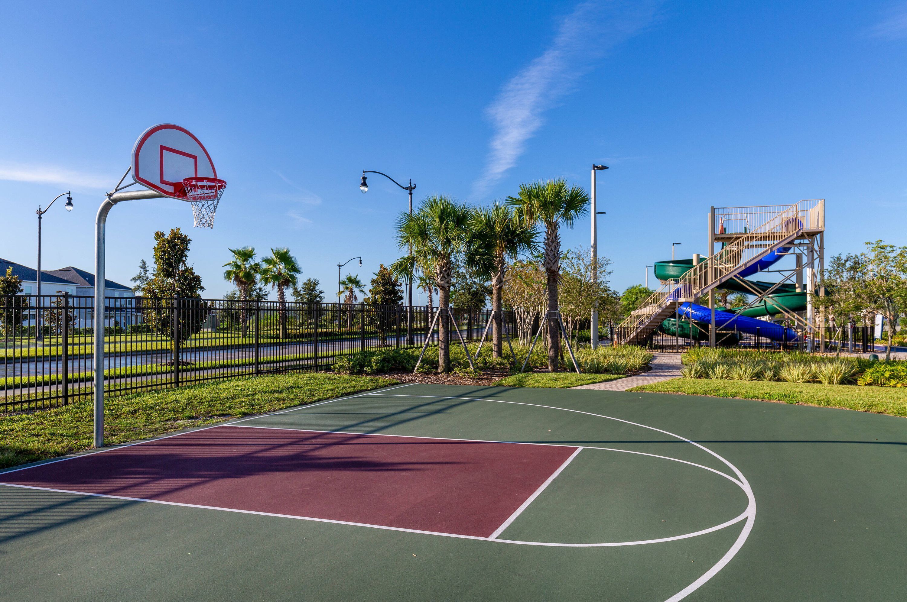 An outdoor basketball court with a green and red playing surface, surrounded by a fence, palm trees, and a water slide structure.