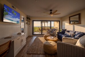 A living room with a television, a ceiling fan, a sofa, a coffee table, and a view of the ocean through large sliding doors.