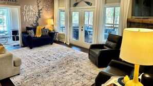 Een woonkamer met een donkerblauwe bank, een beige fauteuil, een kamerkleed met dessin, een plafondventilator en een groot raam met gordijnen.